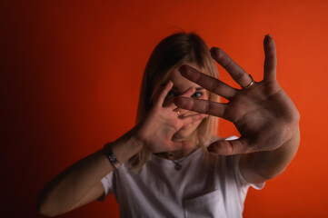 A young woman in a white t-shirt covers her face with her hands against a vibrant orange background. Concept of fear, privacy, rejection, or social anxiety