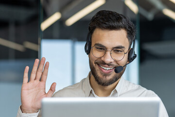 Support service worker with headset and laptop inside office at workplace. Man waving hand in...