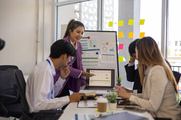 A group of people are sitting around a table with a laptop, a calculator, and a stack of papers. They are discussing business matters and looking at graphs and charts
