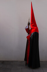 A penitent wearing a tall hood and red tunic stands against a white wall, his covered face embodying the devotion and mystery of Holy Week