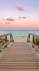 Obraz premium A serene wooden boardwalk leading to a tranquil beach at sunset. The sky is painted in soft pastel colors, with fluffy clouds scattered above. The calm ocean waves gently lap at the shore.