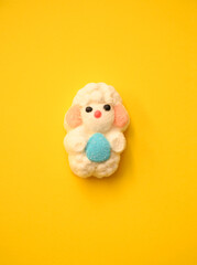 Adorable marshmallow lamb holding a blue Easter egg on a bright yellow background. Perfect for festive decorations, greeting cards, and holiday-themed visuals.