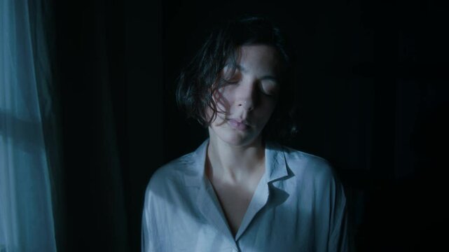 Close-up shot of young woman with dark short hair sleepwalking at night near window in her bedroom