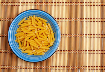 Raw penne pasta in a blue bowl on bamboo mat
