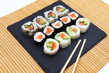 Sushi rolls served on a slate and wooden sushi mat with two chopsticks . Assortment of sushi with salmon , avocado, cucumber and sesame seeds.