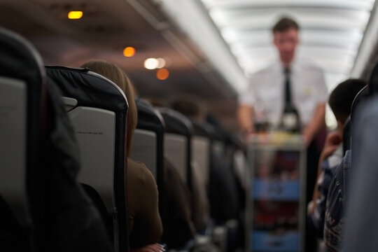 A male steward who distributes food and drinks on board the aircraft. The passengers are sitting in their seats and watching. View along the row. High quality photo