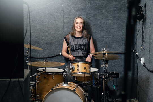 Smiling female drummer sitting behind drum set in music studio. Positive vibe, passion for music, and creativity in rehearsal environment.