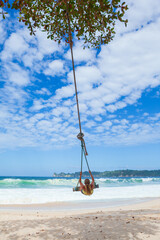 Happy girl have fun swinging high in mid air. Flying up upside down on rope swing on sea beach. Travel adventure on paradise tropical island. Family lifestyle, activity on summer vacation with kids.