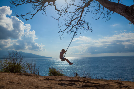 Happy girl have fun swinging high in mid air. Flying up upside down on rope swing on sea beach. Travel adventure on paradise tropical island. Family lifestyle, activity on summer vacation with kids.