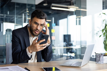 Upset and disappointed man with phone in hands inside office at workplace, businessman reading bad...