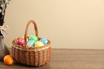 Many decorated Easter eggs in wicker basket and vase with willow branches on wooden table against beige background