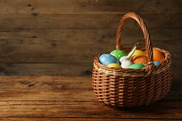 Beautiful decorated Easter eggs and bunny figure in wicker basket on wooden table, space for text