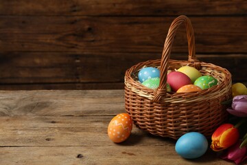 Easter eggs in wicker basket and beautiful tulips on wooden table, space for text