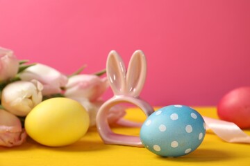Easter eggs, beautiful tulips and bunny figure on yellow wooden table against pink background, closeup