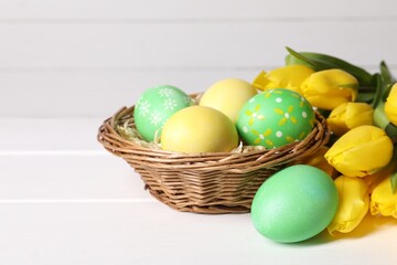 Easter eggs in wicker basket and beautiful tulips on white wooden table, closeup. Space for text