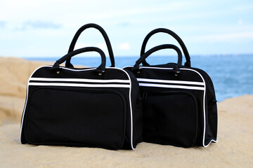 Tourists who have just arrived at the coast place two stylish black bags on the rocky shore against the backdrop of blue sky, sea and shore.