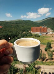 A mug of hot coffee on a balcony with a view of the mountains and the hotel is held by a man's hand