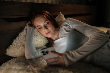 Fototapeta premium Serene redhead young woman lying in bed at night, using smartphone happy looking to screen, enjoying leisure time in cozy bedroom, illuminated by phone screen. Concept of mobile addiction and insomnia