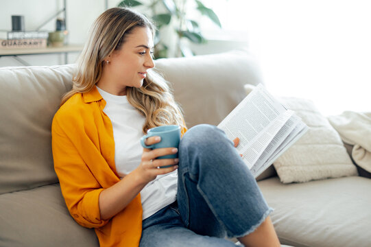 Beautiful calm caucasian woman in casual clothes, sitting on a cozy sofa in a living room, reading a book while holding a cup of tea, relaxed and smiling spending weekend time at home