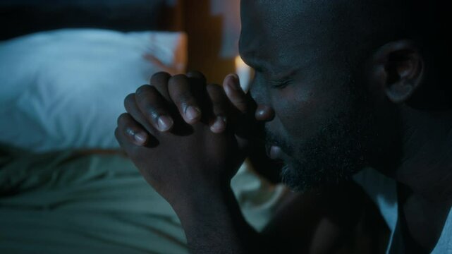 Close-up shot of African American man saying prayers with his eyes closed, clasping hands and kneeling by bed before going to sleep
