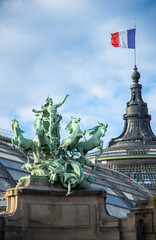 A dramatic Quadriga sculpture dominates the foreground, set against a classic Parisian architectural backdrop. The French flag waves proudly in the sky on a sunny, summer day in Paris, France.