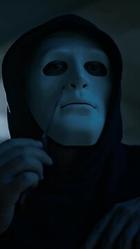 Vertical lowkey closeup portrait of anonymous masked man in hood looking at camera while touching and checking sharp knife, passing it over face and neck