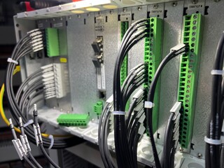 Industrial electrical wiring installation. A technician connects black cables to green terminal blocks inside a control panel. Tools like screwdrivers and a flashlight are on top.