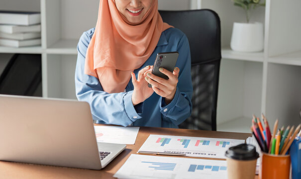 Muslim woman in hijab using smartphone while working at a desk with financial documents. - Powered by Adobe