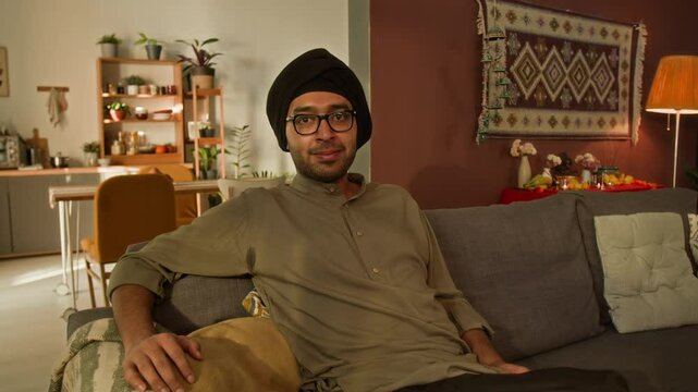 Medium warm light portrait of pleasantly smiling Indian man in glasses, traditional black turban and khaki kurta sitting on couch at home and looking at camera
