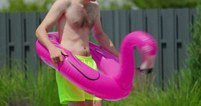 Shirtless man in neon swim shorts dancing joyfully while holding a pink inflatable flamingo pool float near a swimming pool on a hot summer day.
