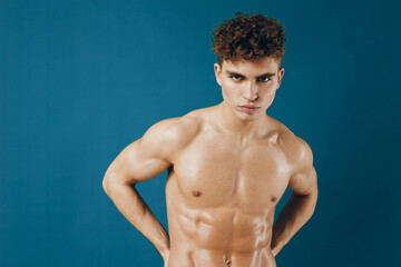 Fototapeta premium Confident young man with curly hair posing shirtless against a teal background, showcasing his athletic physique. Fitness and health concept.
