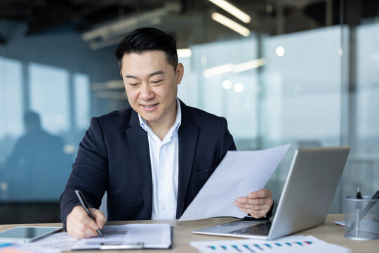Concentrated young Asian man in business suit sitting in office at desk with laptop, working with paper work and signing documents