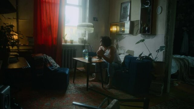 Full shot of exhausted and indifferent man in undershirt and jeans sitting on sofa in old shabby apartment, spending meaningless days alone, questioning decisions of past