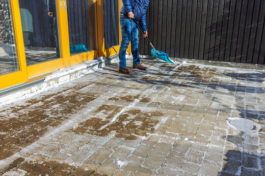Person clearing snow from brick patio with blue plastic shovel during sunny winter afternoon. Sweden.