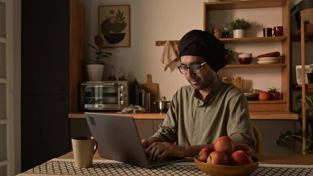 Medium warm light shot of Indian male freelance worker in traditional turban and kurta sitting at dining table in kitchen and coding on laptop