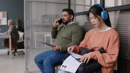 Side shot of diverse visa applicants - bearded Biracial man talking on phone and young Asian lady in headphones - sitting in waiting room before being interviewed in embassy