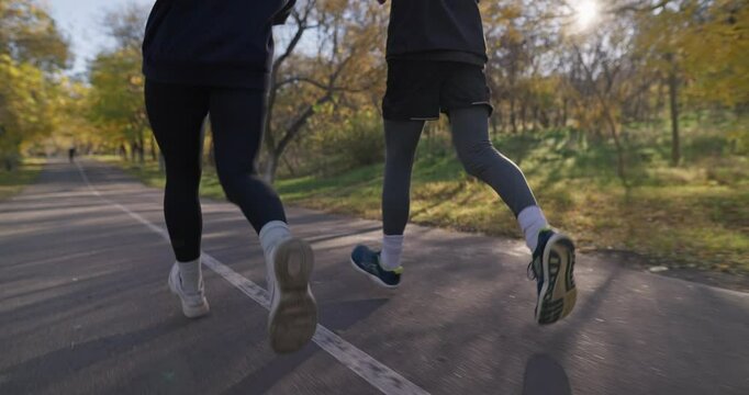 Close up a girl and a guy in black sportswear and sneakers run next to each other along the alley while jogging in the morning in the park