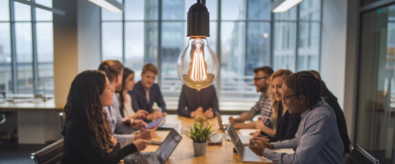 A bright lightbulb suspended above a collaborative meeting where professionals are working together, symbolizing creativity, innovation, and problem-solving in a business environment.