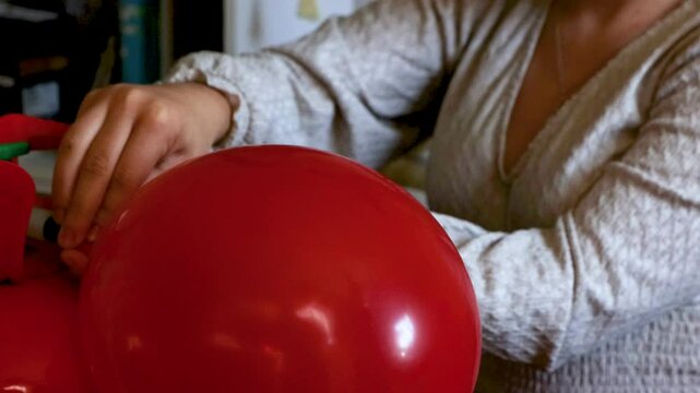 Young woman inflating abd tying a red balloon using a helium tank at home, preparing decorations for a festive event  or birthday celebration in a cozy home environment. 

