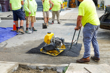 Worker is using a vibratory plate compactor to tamper down newly pored asphalt used to fill a hole in a street