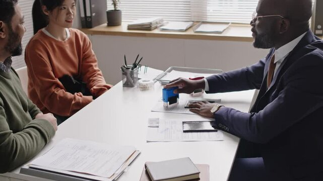 Side shot of smiling African American male visa officer stamping documents while approving visas for diverse spouses during interview in consulate