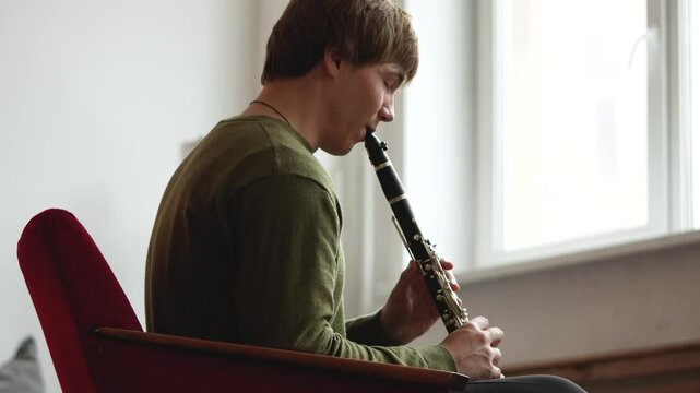 Clarinet player. Concentrated clarinetist playing flute woodwind music instrument sitting on armchair at home. Musical instruments. Man plays the oboe in living room. Relaxed mental hobby concept