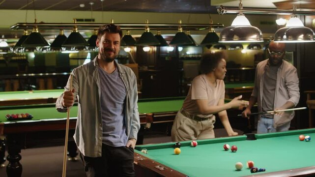 Full medium portrait of young bearded Caucasian man posing by baize table in billiard club with cue stick in hand while playing with diverse friends in evening, look at camera