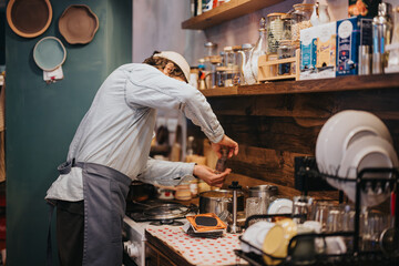 A chef adds seasoning to a dish while cooking in a cozy and organized kitchen, with various cooking utensils and ingredients arranged on the countertop and shelves.