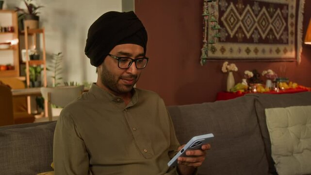Medium portrait of young adult Indian man in glasses, traditional turban and kurta, sitting on sofa and texting messages on phone, looking at camera, hindu altar in background