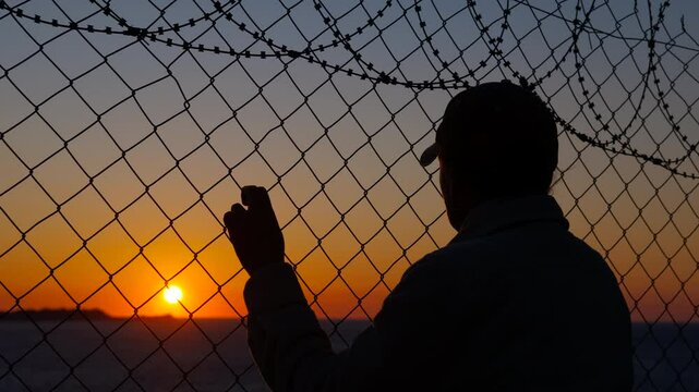 Refugee at sunset near the border. Silhouette of a refugee woman in hopes of a better life clinging to barbed wire against the backdrop of a sea sunset