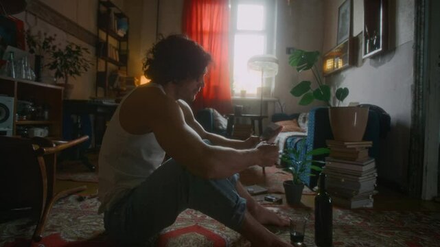 Full side shot of unhappy Caucasian man in undershirt and jeans sitting on floor in messy flat, thinking about breakup with ex-girlfriend and looking at photograph, missing her, feeling lonely
