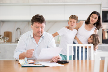 Frustrated man is sitting in kitchen and carefully reading loan agreement. Sympathetic family wife stands behind her husband, hugs stressed children and asks about problems and reasons for bad mood