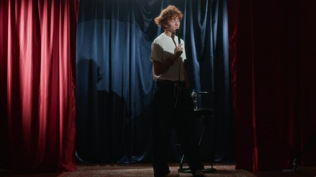 Full low key shot of male red-haired stand-up comedian in white t-shirt casually walking around small stage with red and blue curtains and joking into microphone
