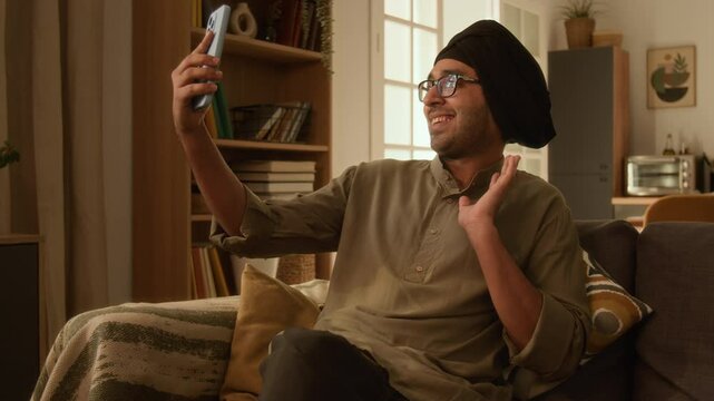 Medium long shot of Indian man with stubble, wearing glasses and turban sitting on comfy couch in living room and video calling friends or family on smartphone